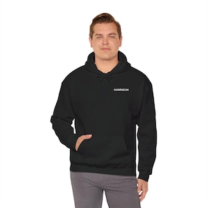 May include: A black hooded sweatshirt with the name "HARRISON" printed in white on the upper chest. The sweatshirt has a front pocket and a drawstring hood. The person is wearing gray pants.
