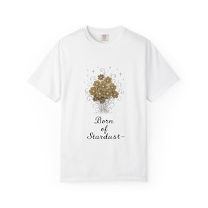 May include: White t-shirt featuring a floral design in shades of gold and brown, with the text "Born of Stardust" in a script font. The design includes stars and swirls. The shirt has a crew neck and short sleeves.