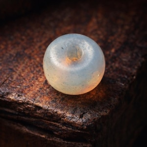 May include: A translucent, donut-shaped bead with a central hole, resting on a dark, textured wooden surface. The bead has a soft, pearly appearance, with subtle color variations and a smooth, rounded form. The lighting highlights its delicate details.
