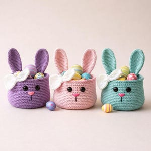 Può includere: Tre cestini a forma di coniglietto all'uncinetto in viola, rosa e verde acqua, ciascuno pieno di uova di Pasqua colorate. I cestini hanno orecchie da coniglio, un fiocco bianco e un naso rosa. Alcune uova decorative sono sparse.