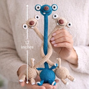 Puede incluir: Un juguete de ganchillo hecho a mano, de aproximadamente 30 cm de altura. El juguete presenta una cabeza y un cuerpo azules, con brazos y piernas beige. La criatura tiene ojos grandes y redondos y una cara sonriente. El diseño es juguetón y colorido, adecuado para un juguete infantil o la decoración de una guardería.