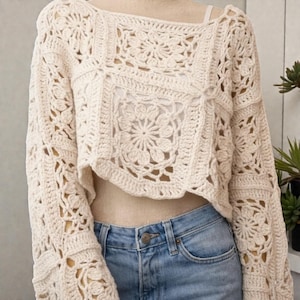 May include: Cream-colored, cropped crochet sweater with floral and square patterns. The long-sleeved top features an openwork design and is paired with blue jeans. The sweater is made of a soft, textured yarn.
