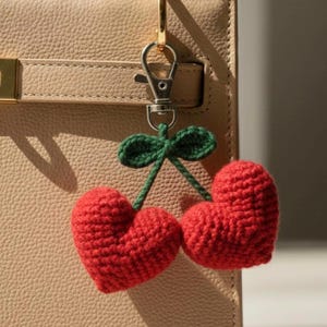 May include: A handmade red crochet cherry charm with green leaves and stem, attached to a silver-colored clasp. The charm is hanging from a beige leather handbag.