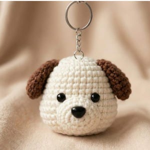 May include: A handmade crochet dog keychain. The dog's head is white with brown ears, black eyes, and a black nose. A silver-colored metal keyring and chain are attached to the top of the dog's head. The background is a soft, neutral color.