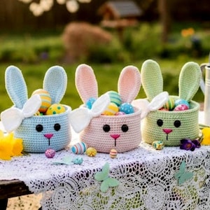 May include: Three handmade crochet Easter bunny baskets in blue, pink, and green, each filled with colorful Easter eggs. The baskets have bunny ears and faces, and are set on a white lace tablecloth.