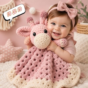 May include: A crocheted pink giraffe lovey with a cream-coloured snout and a matching blanket. The blanket measures approximately 48 x 48 cm. The image also includes a baby wearing a pink headband with a bow.