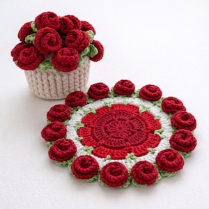 Pode incluir: Um conjunto de crochê feito à mão com uma cesta de rosas vermelhas e um porta-copos a condizer. O porta-copos é redondo, com um design floral vermelho central, rodeado por detalhes de rosas brancas, verdes e vermelhas. A cesta é bege com detalhes verdes.