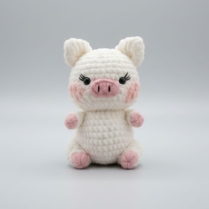 May include: A handmade white crochet pig with pink accents. The pig has a pink snout, blush-colored cheeks, and small black eyes with eyelashes. The pig is sitting upright, with its legs and feet in pink.