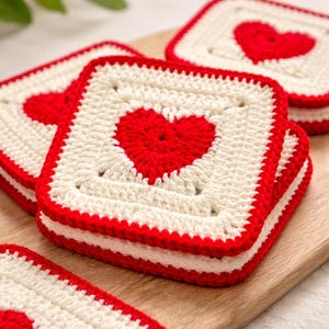 May include: A stack of square, crocheted coasters. Each coaster features a red heart on a cream-colored background, framed by a red border. The coasters are stacked on a wooden surface, showcasing the handmade quality and design.