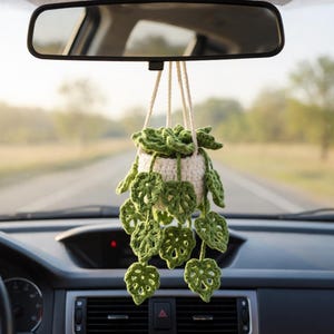 May include: A crocheted car air freshener featuring a small white pot with green leaves. The leaves are a vibrant green color and hang down from the pot. The air freshener is suspended from the rearview mirror by white cords. The car's dashboard is visible in the foreground.