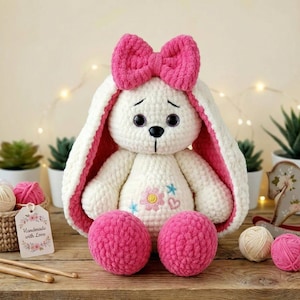 Op de afbeelding: Een handgemaakt wit gehaakt konijn met een grote roze strik, roze binnenoren en roze pomponvoeten. Het konijn heeft geborduurde gezichtskenmerken en een bloemenontwerp op de borst. Een label leest "Handmade with Love".