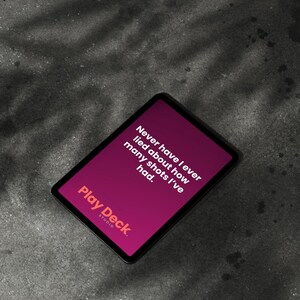 以下が含まれることがあります： 「Play Deck Studio」のゲームカード。マゼンタ色の背景に「Never have I ever lied about how many shots I've had.」のテキスト。カードは黒い縁取りで、灰色の表面に置かれています。