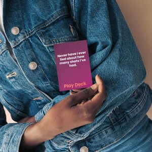 以下が含まれることがあります： 「Never have I ever lied about how many shots I've had」と書かれた紫色のPlay Deckカードを持つ人。その人はデニムジャケットを着ています。