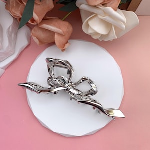 May include: A silver-colored hair claw clip in a bow shape, resting on a white circular platform. The clip has a textured, twisted design. The background features pink and peach-colored artificial flowers.