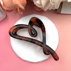 Può includere: Una molletta per capelli a forma di cuore con motivo tartarugato. La clip è marrone con dettagli neri e ha una finitura opaca. È appoggiata su una superficie bianca di forma geometrica, con fiori rosa e crema sullo sfondo.