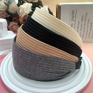 Könnte beinhalten: Ein Stapel von fünf gerippten Haarbändern in verschiedenen Farben: Grau, Beige, Schwarz, Creme und Hellbeige. Die Haarbänder sind auf einem weißen, runden, mehrstufigen Tablett angeordnet. Im Hintergrund ein Blumenstrauß und eine Papiertüte.