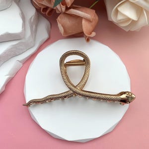 Könnte beinhalten: Goldfarbene Haarspange in Schlangenform. Der Clip ist mit einem strukturierten Muster und kleinen, runden Akzenten versehen. Der Schlangenkopf befindet sich an einem Ende. Der Clip liegt auf einer weißen, geometrischen Oberfläche.