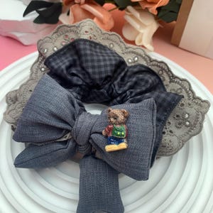 Puede incluir: Un lazo para el cabello de tela gris con un pequeño dije de oso de peluche. El lazo está superpuesto a una diadema con ribete de encaje gris y una tela de cuadros grises oscuros. Rosas color melocotón en el fondo.