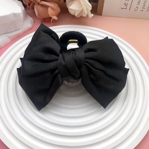 May include: A large black bow hair clip made of a silky fabric. The bow is centered on a black clip with a gold-colored accent. The hair accessory is displayed on a white, ridged plate.