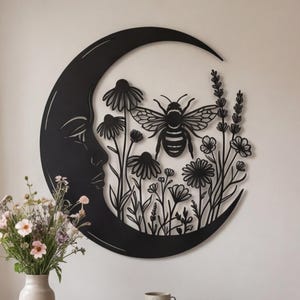 May include: A black metal wall art piece shaped like a crescent moon with a human face profile, featuring a large bee surrounded by various wildflowers and stems. The design is intricate, with cut-out details forming the floral and insect shapes.