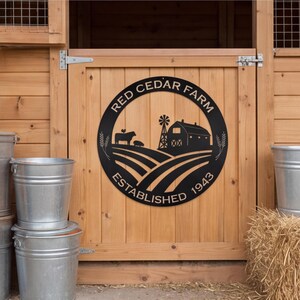Puede incluir: Un letrero circular de metal negro con una escena de granja que incluye un granero, un molino de viento, una vaca y un cerdo. El texto dice RED CEDAR FARM y ESTABLISHED 1943. El diseño incluye espigas de trigo y líneas curvas de campos.