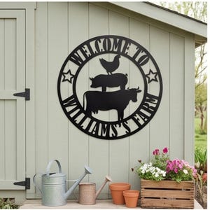 Może przedstawiać: Okrągły czarny metalowy znak ścienny z sylwetką kurczaka, świni i krowy ułożonych pionowo. Napis "WELCOME TO WILLIAM'S FARM" jest wycięty wzdłuż okrągłej krawędzi i ozdobiony dwiema pięcioramiennymi gwiazdami po obu stronach.