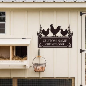 Puede incluir: Un letrero metálico marrón oscuro para un gallinero, con siluetas de un gallo y dos gallinas sobre el texto "CUSTOM NAME CHICKEN COOP". El letrero está montado en una pared de madera color crema, con una cesta de alambre con huevos colgando debajo.