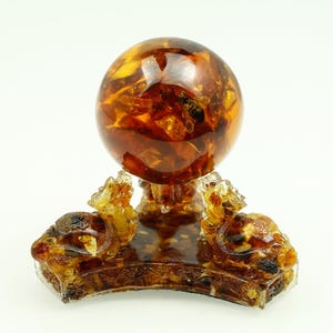 Puede incluir: Una esfera decorativa de ámbar, que muestra una mezcla de tonos dorados y marrones. La esfera descansa sobre una base de ámbar ornamentada con tallas detalladas. Un artículo decorativo único.
