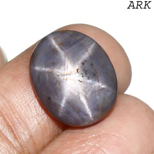 May include: A polished, oval-shaped, gray-blue star sapphire gemstone. The stone displays a six-rayed star pattern across its surface. The word "ARK" is printed in black, upper-case letters in the upper right corner.