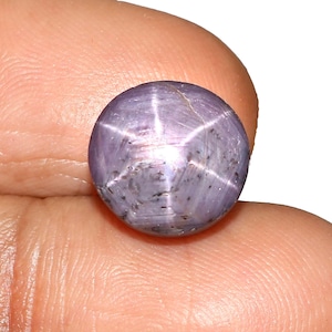 May include: A spherical, lilac-hued gemstone displaying a six-rayed star pattern. The gem is held between fingers, highlighting its polished surface and the star effect from light refraction. The text "ARK" is visible in the upper right corner.