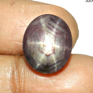 May include: Oval-shaped star ruby gemstone. The gem has a deep purple colour with a six-rayed star pattern across its surface. The stone is held between fingers, showcasing its polished, reflective facets.