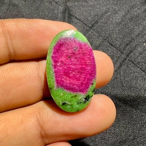 Puede incluir: Una piedra preciosa en forma de cabujón ovalado con un centro rosa magenta vibrante rodeado por un borde verde lima brillante. La piedra muestra una superficie lisa y pulida con variaciones de color naturales y motas oscuras en el área verde.
