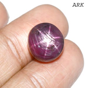 Puede incluir: Una gema de rubí pulida, tallada en cabujón, con efecto estrella. La gema es de un rojo intenso, con un patrón de estrella de seis rayos en su superficie. La palabra "ARK" es visible en la esquina superior derecha.