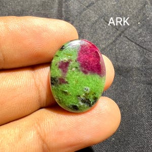 May include: An oval-shaped cabochon gemstone featuring a vibrant mix of bright green zoisite and deep magenta ruby inclusions, with small black mineral flecks throughout. The stone is polished to a smooth finish and displays the text ARK.