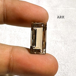 May include: A rectangular, step-cut smoky quartz gemstone with a translucent, light brown hue and faceted edges. The stone features a clear, polished surface and is marked with the text ARK in the upper right corner of the frame.