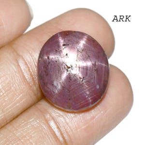 May include: A polished, oval-shaped, star ruby gemstone. The ruby is a deep pinkish-red color with a six-rayed star effect across its surface. The word "ARK" is printed in black in the upper right corner.