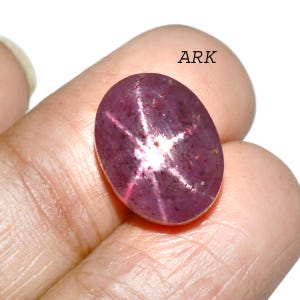 May include: Oval-shaped, purplish-red gemstone with a six-rayed star pattern. The gem has a polished surface and a white star in the center. The text "ARK" is visible in the upper right corner.