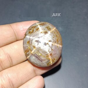 May include: Oval-shaped gemstone with a star-like pattern. The stone has a mix of brown, gold, and gray hues, with a prominent star effect visible across its surface. The word "ARK" is visible in the upper right corner.