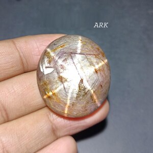 May include: A polished, oval-shaped gemstone with a star-like pattern. The stone has a mix of brown, tan, and white hues, with a prominent six-rayed star design. The word "ARK" is visible in the upper right corner. The gemstone is held in a hand.
