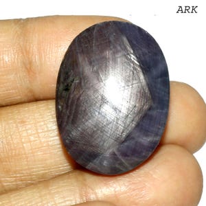 May include: An oval-shaped, dark purple gemstone with a star-like effect. The stone has a polished surface with visible lines and a subtle sheen. The gemstone is held in a hand, showcasing its size and color.