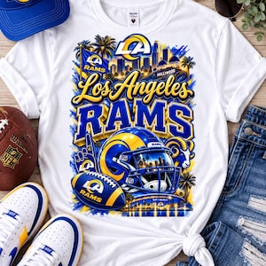 Pode incluir: Uma t-shirt branca de manga curta com um gráfico dos Los Angeles Rams, incluindo um capacete de futebol americano, uma bola, palmeiras e o horizonte da cidade. O texto diz Los Angeles Rams e Hollywood nas cores azul e amarelo.