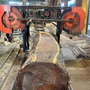 May include: A large, dark brown log is being cut by an industrial band saw with orange and black components. The log's end grain is visible, showing its natural wood grain. The saw is in a workshop setting, with the text "Quali EpoxyTablesCo" visible.