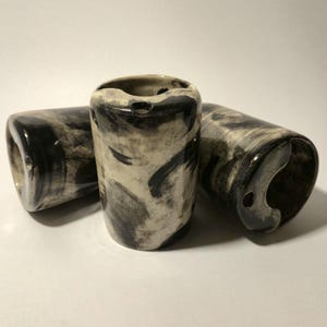 Puede incluir: Tres vasos de chupito de cerámica con forma cilíndrica. Los vasos son de color blanquecino con un diseño veteado negro. Cada vaso tiene un pequeño agujero cerca de la parte superior. Los vasos de chupito están colocados sobre una superficie blanca.