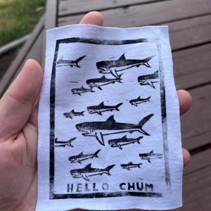 May include: A white fabric patch featuring a black print of various shark illustrations. The phrase "HELLO CHUM" is printed in bold, capital letters at the bottom. The patch is held in a hand.