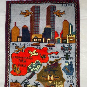 May include: A rectangular woven rug featuring a gray background with a skyline of two tall buildings and airplanes at the top. The lower section displays a map of Afghanistan and Iraq with military vehicles, helicopters, and text including 9-11-01.