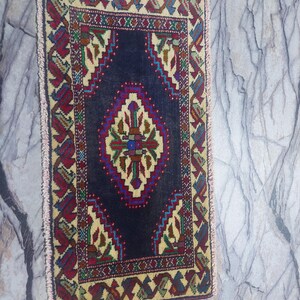May include: A rectangular, hand-woven rug with a dark blue central field and a cream-coloured border. The border features repeating geometric patterns in red, green, and blue. The rug has a central diamond-shaped design with intricate details and fringe on the edges.
