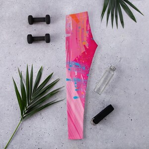 Könnte beinhalten: Rosa und orangefarbene Leggings mit abstraktem Muster und blauen Farbspritzern. Die Leggings liegen neben zwei schwarzen Kurzhanteln, einer durchsichtigen Wasserflasche und einer schwarzen Handschlaufe. Ein grünes Palmenblatt ist ebenfalls im Bild.