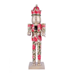 May include: A decorative wooden nutcracker figurine in a standing pose. The figurine is painted silver with red and green floral accents. It has a crown, drum, and drumsticks. The nutcracker stands on a rectangular base.