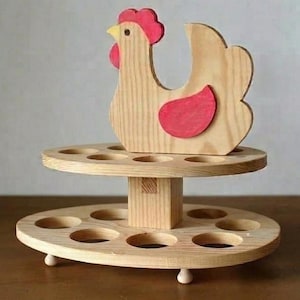 Könnte beinhalten: Zweistöckiger Eierhalter aus Holz mit dekorativem Huhn. Der Ständer hat kreisförmige Aussparungen für Eier. Das Huhn hat rote Akzente auf Kamm und Kehllappen. Der Ständer ist aus hellem Holz gefertigt.