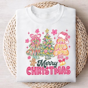 Pode incluir: Camisola branca com um design colorido de árvore de Natal. O gráfico inclui três árvores estilizadas, flores cor-de-rosa, um homem de gengibre e o texto "Merry Christmas" em rosa e verde. O design tem uma estética retro e caprichosa.
